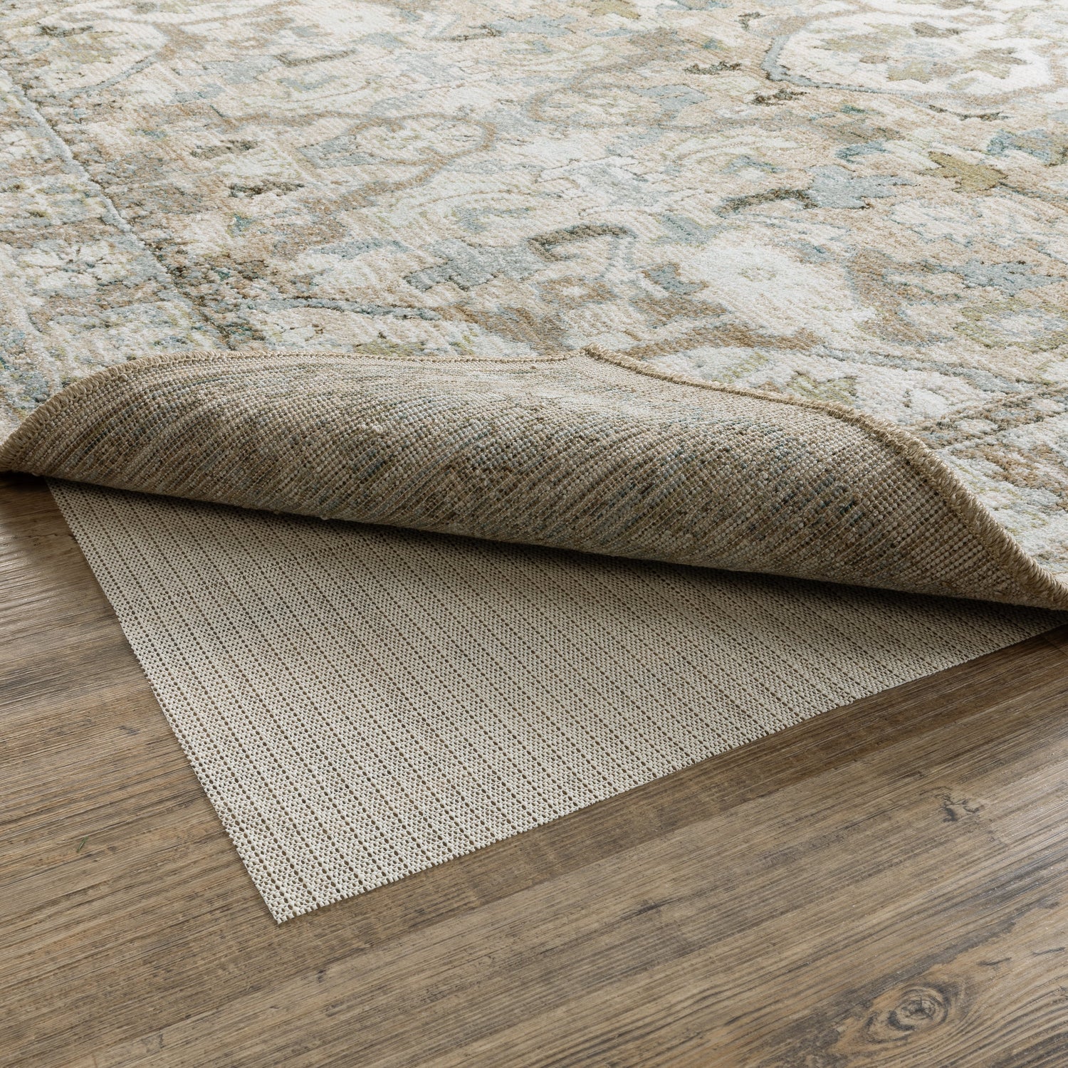 A patterned beige-and-pale-blue area rug is partially lifted, revealing a textured beige non-slip rug pad beneath on a light wood floor.
