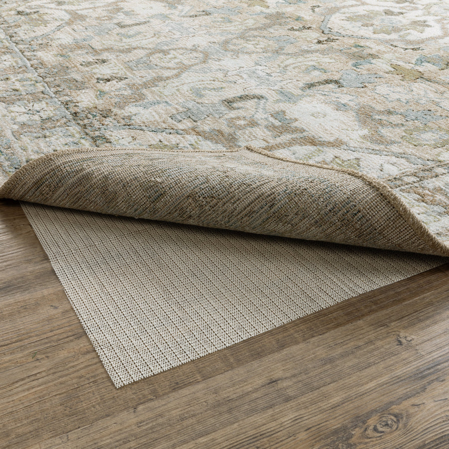 A patterned beige-and-pale-blue area rug is partially lifted, revealing a textured beige non-slip rug pad beneath on a light wood floor.