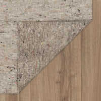 Felt rug pad corner folded back, revealing light brown wood-plank flooring beneath; pad displays multicolored recycled fibers and a textured, compressed surface against the wooden floor.
