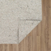 A beige felt rug pad is folded back, revealing brown wood-grain laminate flooring beneath; fibers and small specks dot the padding against the smooth plank surface in a corner layout.