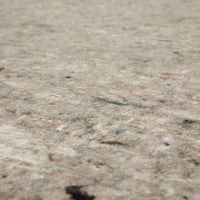 Beige felt-like rug pad lies flat, showing mottled fibers and small colored specks; close-up low-angle view emphasizes soft, slightly fuzzy texture across an indoor surface.