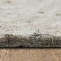 A thick fibrous felt rug pad rests on a wooden surface, its frayed edge and loose hairs visible in a close-up macro shot with a soft, blurred neutral background.
