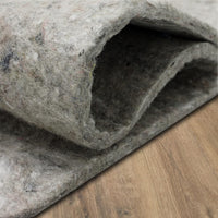 A folded gray felt rug pad lies partially rolled open on a hardwood floor, revealing dense, fuzzy fibers and layered thickness.