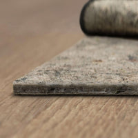 Rectangular felt rug or carpet pad lies flat on a wooden floor, its fibrous, worn surface shown in close-up while a rolled carpet sample blurs in the background.