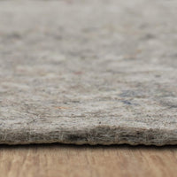 A thick, fibrous gray felt rug pad lies flat, its textured edge visible in close-up, resting on a wooden surface with a soft, out-of-focus pale background.