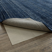 Blue area rug folded back to show a beige non-slip rug pad with a woven grip surface on a wood floor