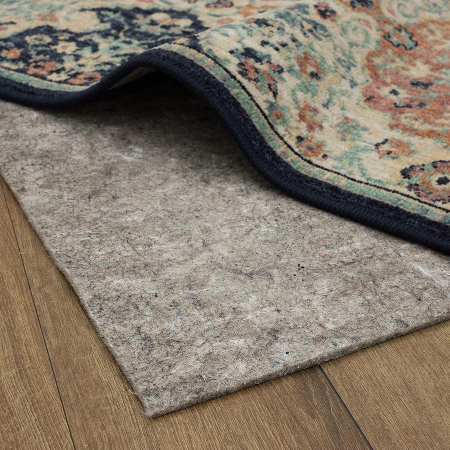 A patterned area rug peels up at one corner, revealing a gray felt rug pad beneath; ornate teal, rust, and cream design with dark blue binding sits atop wood flooring.