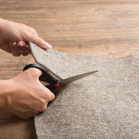 Hands hold black-handled scissors cutting a gray rug pad.