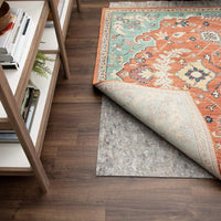Patterned rug corner folded back to reveal a gray rug under pad on dark hardwood, beside a wooden shelf holding books and a small potted fern.